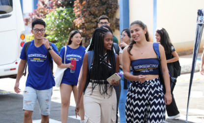 Na imagem, estão, da esquerda para a direita, um estudante oriental, de camisa azul e short jeans; uma estudante preta de camisa azul e calça bege, com cabelos trançados; uma estudante branca de cropped azul com escrito "biomedicina UNIFAL-MG" e uma calça com ziguezague desenhado de preto com fundo branco. Atrás destes estudantes há outros estudantes cuja identificação fica restrita pelo desfoque. Os estudantes estão entrando na Universidade para cursarem o primeiro semestre de 2024, interagindo, sorrindo e em um momento descontraído. Fim da Descrição.