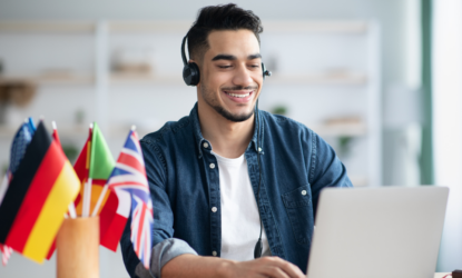 Na imagem, há um jovem branco, de cabelo preto e barba, blusa branca e camisa jeans, fones nos ouvidos, em frente a um notebook. Ao lado do rapaz, há um copo com bandeirinhas de diversos países. Ele está em, aparentemente, uma sala de aula, desfocada de fundo. Fim da descrição.