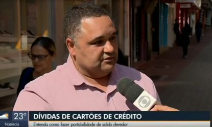 Na imagem, o professor Marçal está em frente ao repórter, falando ao microfone, vestido com uma camisa rosa de botão. Na parte inferior da imagem há um escrito branco sobre uma faixa azul "DÍVIDAS DE CARTÕES DE CRÉDITO". Abaixo, escrito de azul sobre uma faixa branca "ENTENDA COMO FAZER PORTABILIDADE DE SALDO DEVEDOR". FIm da descrição.