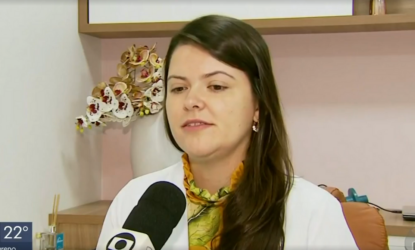 Na imagem, a professora Maria Olivia está olhadno para frente, em frente ao microfone do repórter, vestida de jaleco branco, em uma sala de consultório. Fim de descrição.