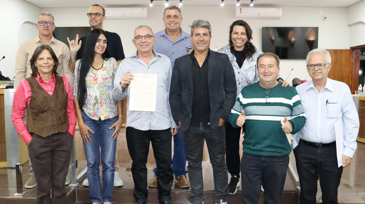 A foto mostra dez pessoas - entre os quais o homenageado ao meio, segurando a moção recebida, e os vereadores da Casa Legislativa de Alfenas - em pose olhando para o fotógrafo.