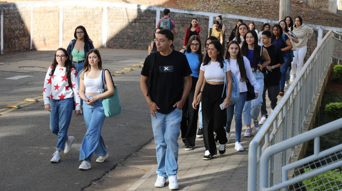 Na imagem há, aproximadamente, 23 estudantes da UNIFAL-MG caminhando na Universidade, durante à Acolhida aos Calouros 2024/2. Todos eles estão de calça, tênis e camiseta. Há uma diversidade étnica presente na imagem. Fim da descrição.