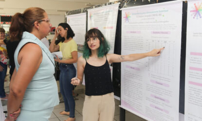 Na imagem, estão duas mulheres. À esquerda, está uma mulher ouvindo a apresentação de uma estudante, que está à direita, apontando para o banner. A espectadora está com os braços cruzados para trás e veste um conjunto de blusa e calça azuis. A estudante, localizada à direita da imagem, tem cabelos azuis e veste uma blusa preta e calça marrom. Fim da descrição.