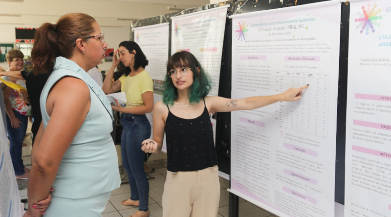 Na imagem, estão duas mulheres. À esquerda, está uma mulher ouvindo a apresentação de uma estudante, que está à direita, apontando para o banner. A espectadora está com os braços cruzados para trás e veste um conjunto de blusa e calça azuis. A estudante, localizada à direita da imagem, tem cabelos azuis e veste uma blusa preta e calça marrom. Fim da descrição.