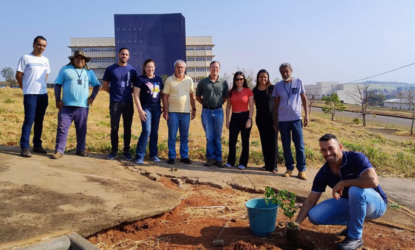 Na imagem, estão posicionados de corpo inteiro servidores da Universidade Federal de Alfenas, em um gramado, com um prédio azul de fundo. Logo na parte central direita da imagem, há um servidor terceirizado, de camiseta azul e calça jeans, posicionado de modo agachado, ao lado de uma muda prestes a ser plantada. Todos olham para a câmera e sorriem. Fim da descrição.