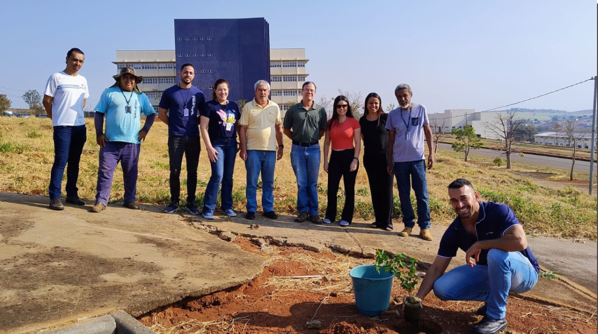 Na imagem, estão posicionados de corpo inteiro servidores da Universidade Federal de Alfenas, em um gramado, com um prédio azul de fundo. Logo na parte central direita da imagem, há um servidor terceirizado, de camiseta azul e calça jeans, posicionado de modo agachado, ao lado de uma muda prestes a ser plantada. Todos olham para a câmera e sorriem. Fim da descrição.