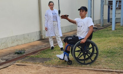 A imagem é uma foto A imagem mostra uma foto de um paratleta em uma cadeira de rodas realizando um movimento com o braço estendido, participando de um exercício físico ao ar livre. Ele está vestindo uma camiseta branca e boné preto. Ao lado, uma moça com jaleco branco, observa a ação. Eles estão próximos de uma área com areia, em um ambiente externo, ao lado de um prédio.