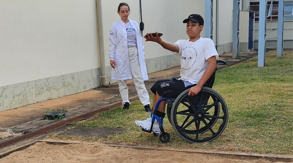 A imagem é uma foto A imagem mostra uma foto de um paratleta em uma cadeira de rodas realizando um movimento com o braço estendido, participando de um exercício físico ao ar livre. Ele está vestindo uma camiseta branca e boné preto. Ao lado, uma moça com jaleco branco, observa a ação. Eles estão próximos de uma área com areia, em um ambiente externo, ao lado de um prédio.