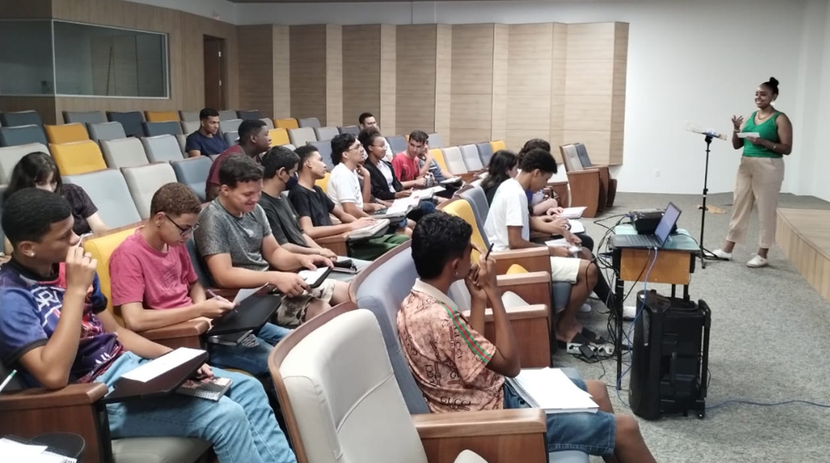 A imagem mostra um grupo de estudantes sentados em um auditório, prestando atenção e fazendo anotações enquanto uma das acadêmicas envolvidas no projeto, está de pé à frente da sala, ministrando a oficina.