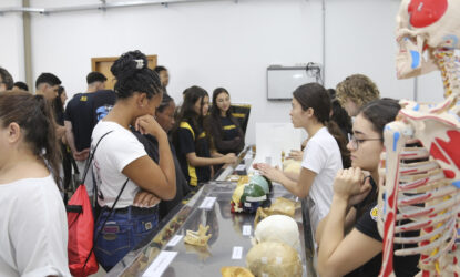 A imagem é uma foto que mostra um grupo de estudantes em um laboratório, interagindo com uma exposição de itens anatômicos, incluindo modelos de crânios e um esqueleto humano. Algumas pessoas estão observando e outras conversando com expositores que estão explicando os materiais expostos.