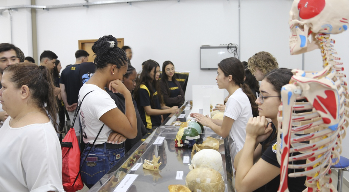 A imagem é uma foto que mostra um grupo de estudantes em um laboratório, interagindo com uma exposição de itens anatômicos, incluindo modelos de crânios e um esqueleto humano. Algumas pessoas estão observando e outras conversando com expositores que estão explicando os materiais expostos.