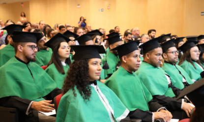 Na imagem, estão diversos estudantes graduados, de diferentes cores e etnias, sentados e enfileirados, vestidos de beca com o manto verde e com os capelos nas cabeças. Fim da descrição.