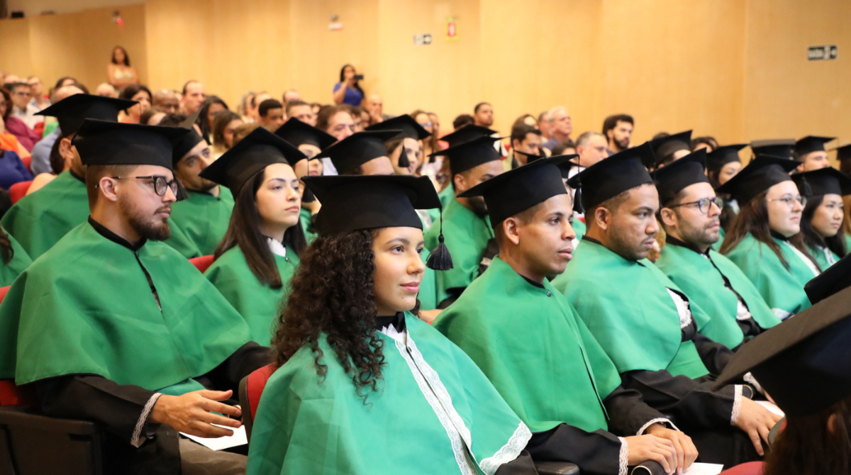 Na imagem, estão diversos estudantes graduados, de diferentes cores e etnias, sentados e enfileirados, vestidos de beca com o manto verde e com os capelos nas cabeças. Fim da descrição.