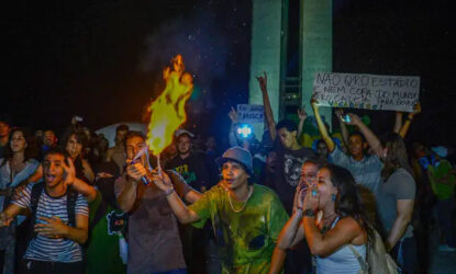 A imagem mostra uma foto de um grupo de manifestantes durante um protesto noturno nas Jornadas 2013. No centro, uma pessoa segura um objeto em chamas, enquanto outras ao redor expressam emoção e participam ativamente. Ao fundo, algumas pessoas seguram cartazes, incluindo um que critica investimentos em estádios e eventos esportivos, refletindo insatisfação com gastos públicos.