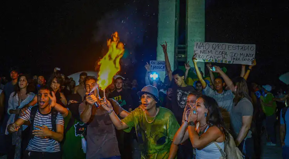 A imagem mostra uma foto de um grupo de manifestantes durante um protesto noturno nas Jornadas 2013. No centro, uma pessoa segura um objeto em chamas, enquanto outras ao redor expressam emoção e participam ativamente. Ao fundo, algumas pessoas seguram cartazes, incluindo um que critica investimentos em estádios e eventos esportivos, refletindo insatisfação com gastos públicos.