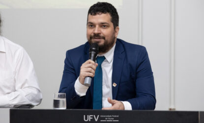 A imagem mostra o professor convidado para o seminário Evandro Rodrigues de Faria. Ele está vestindo um terno azul marinho e gravata azul, segurando um microfone enquanto fala em um ambiente formal. Ele está sentado atrás de uma mesa, onde há um copo de água. À frente da mesa, vê-se o logotipo da Universidade Federal de Viçosa (UFV).