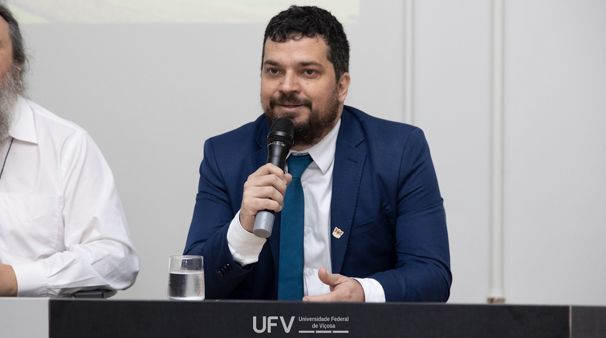 A imagem mostra o professor convidado para o seminário Evandro Rodrigues de Faria. Ele está vestindo um terno azul marinho e gravata azul, segurando um microfone enquanto fala em um ambiente formal. Ele está sentado atrás de uma mesa, onde há um copo de água. À frente da mesa, vê-se o logotipo da Universidade Federal de Viçosa (UFV).