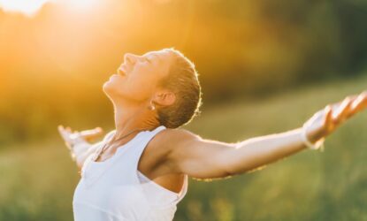 A imagem mostra uma mulher de cabelos curtos e de braços abertos em um campo ao ar livre, sob luz solar intensa, com uma expressão de alegria e serenidade.