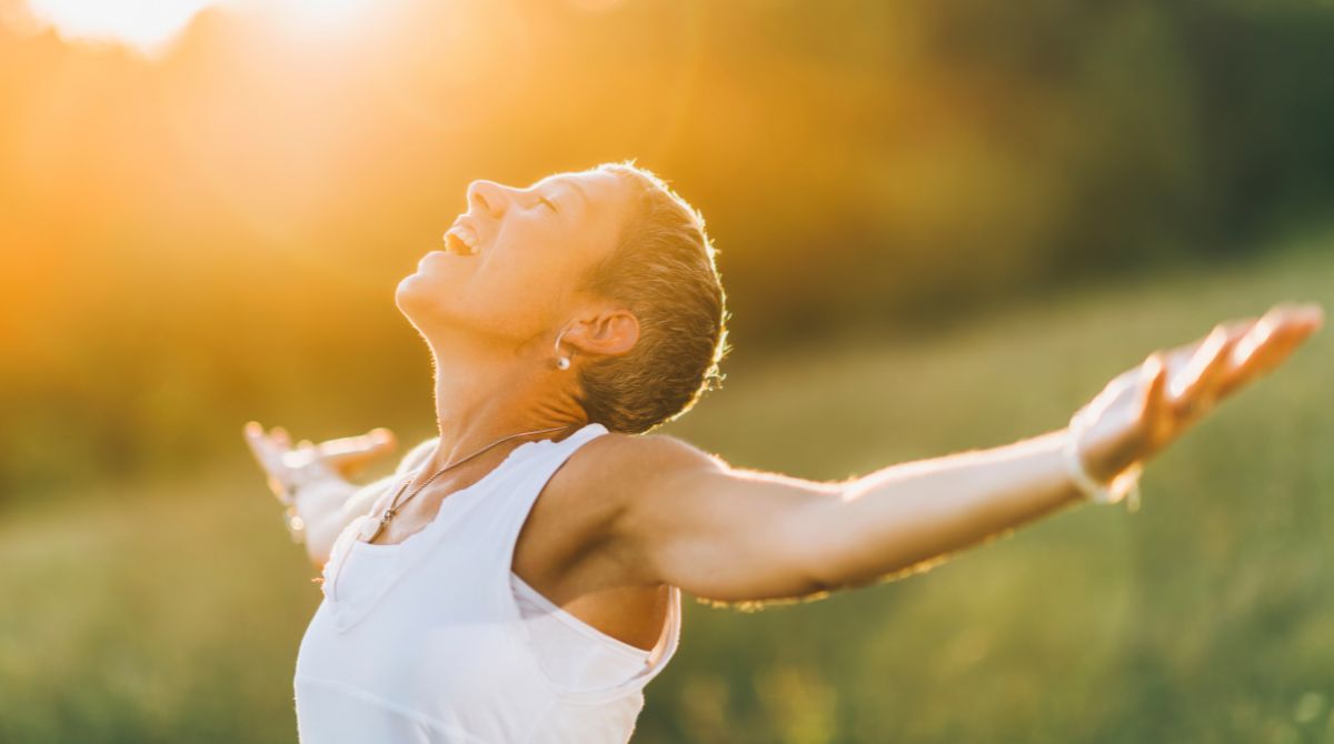 A imagem mostra uma mulher de cabelos curtos e de braços abertos em um campo ao ar livre, sob luz solar intensa, com uma expressão de alegria e serenidade.