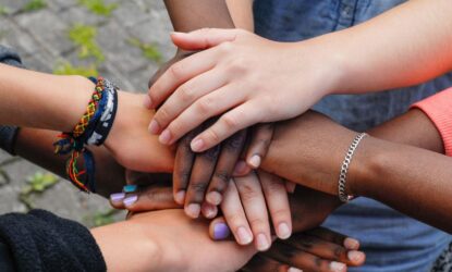 A imagem mostra uma foto de várias mãos de diferentes cores e estilos unidas em um gesto de solidariedade e apoio mútuo. As mãos se sobrepõem, simbolizando diversidade, cooperação e união.