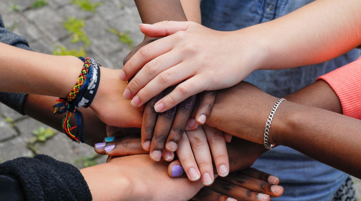 A imagem mostra uma foto de várias mãos de diferentes cores e estilos unidas em um gesto de solidariedade e apoio mútuo. As mãos se sobrepõem, simbolizando diversidade, cooperação e união.