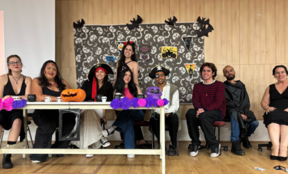 A imagem mostra um grupo de nove pessoas posando para uma foto em uma sala decorada para o Halloween. Elas estão sentadas ou em pé atrás de uma mesa enfeitada com abóboras, pequenos crânios e flores de papel nas cores roxa e lilás. A parede ao fundo tem várias figuras de morcegos, bandeirinhas e pôsteres com temas de Halloween, incluindo imagens de bruxas e outros elementos assustadores. As pessoas na foto estão fantasiadas ou usando acessórios temáticos. Da esquerda para a direita: Primeira pessoa: está usando óculos e um vestido preto, com uma expressão serena. Segunda pessoa: tem um rosto pintado como se estivesse ferido e veste uma blusa preta. Terceira pessoa: usa um chapéu de pirata vermelho e preto, com um sorriso. Quarta pessoa: está com orelhas de gato na cabeça e usa uma camiseta escura. Quinta pessoa (em pé, no centro): está vestida de diabinha com um par de chifres vermelhos e um sorriso no rosto. Sexta pessoa: também usa um chapéu de pirata e um tapa-olho, com uma expressão alegre. Sétima pessoa: veste uma blusa listrada preta e vermelha, com um sorriso descontraído. Oitava pessoa: usa uma capa preta e tem uma expressão mais séria. Nona pessoa: está vestida de preto e sorri para a câmera. A decoração e as fantasias criam um clima festivo e descontraído, ideal para uma celebração de Halloween.
