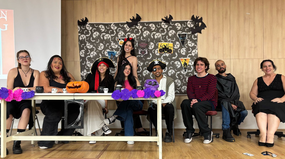 A imagem mostra um grupo de nove pessoas posando para uma foto em uma sala decorada para o Halloween. Elas estão sentadas ou em pé atrás de uma mesa enfeitada com abóboras, pequenos crânios e flores de papel nas cores roxa e lilás. A parede ao fundo tem várias figuras de morcegos, bandeirinhas e pôsteres com temas de Halloween, incluindo imagens de bruxas e outros elementos assustadores. As pessoas na foto estão fantasiadas ou usando acessórios temáticos. Da esquerda para a direita: Primeira pessoa: está usando óculos e um vestido preto, com uma expressão serena. Segunda pessoa: tem um rosto pintado como se estivesse ferido e veste uma blusa preta. Terceira pessoa: usa um chapéu de pirata vermelho e preto, com um sorriso. Quarta pessoa: está com orelhas de gato na cabeça e usa uma camiseta escura. Quinta pessoa (em pé, no centro): está vestida de diabinha com um par de chifres vermelhos e um sorriso no rosto. Sexta pessoa: também usa um chapéu de pirata e um tapa-olho, com uma expressão alegre. Sétima pessoa: veste uma blusa listrada preta e vermelha, com um sorriso descontraído. Oitava pessoa: usa uma capa preta e tem uma expressão mais séria. Nona pessoa: está vestida de preto e sorri para a câmera. A decoração e as fantasias criam um clima festivo e descontraído, ideal para uma celebração de Halloween.