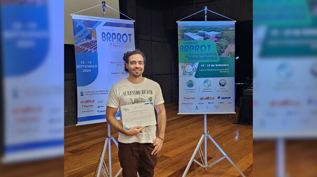 A imagem mostra um homem de pé segurando um certificado, sorrindo para a câmera. Ele está em um ambiente de conferência, com banners ao fundo. Os banners indicam que ele está no "6º Congresso BRPROT + Summit Brasileiro de Metabolômica", realizado entre 16 e 19 de setembro de 2024 no Centro de Difusão Internacional da USP, em São Paulo. No certificado que ele segura, é possível perceber seu reconhecimento no evento. Ele veste uma camiseta clara com a estampa "Surfside Beach" e calça marrom escura.