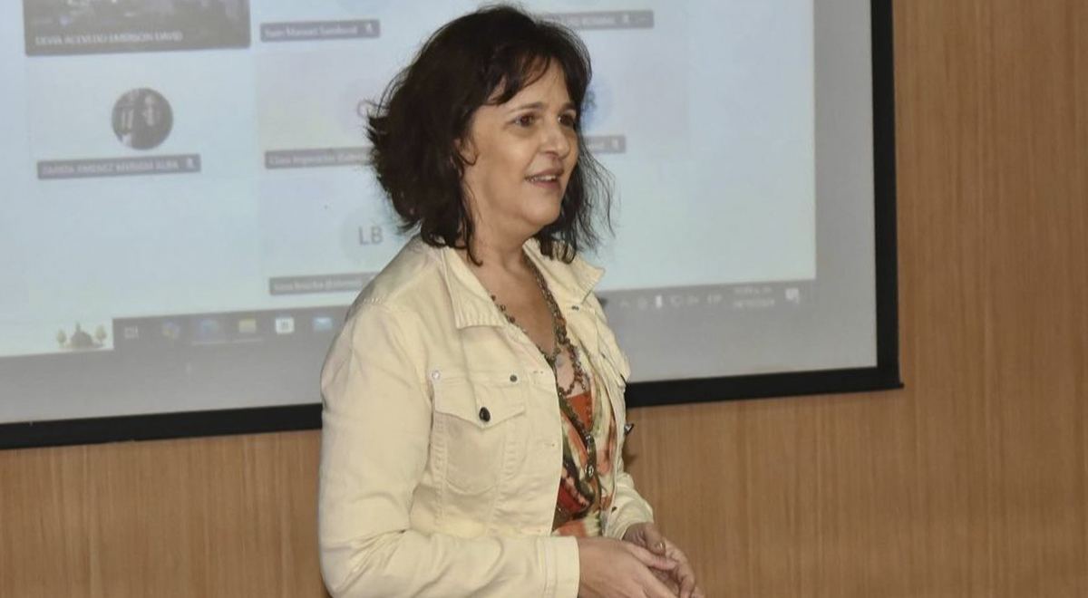 A imagem é uma foto da professora Adriana Imperador durante a apresentação no evento. Ela é uma mulher com cabelos castanhos e curtos, usando uma jaqueta bege e blusa estampada, de pé em frente a uma tela de projeção em ambiente fechado, dando a palestra. Ao fundo, é possível ver partes desfocadas de um slide com nomes e ícones.