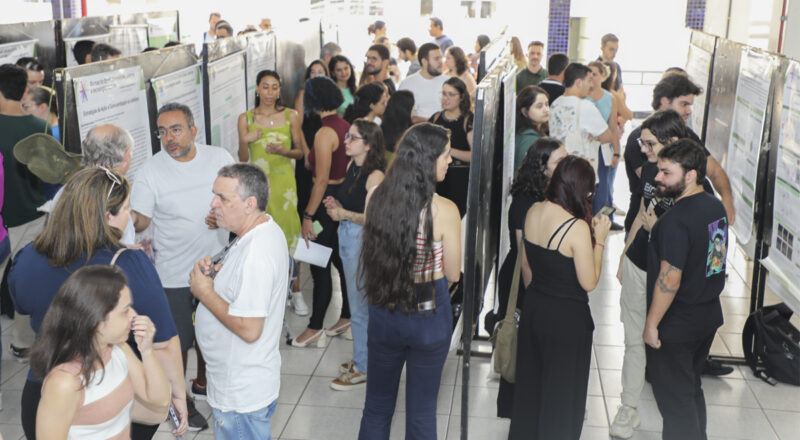 Imagem mostra a foto da apresentação de trabalhos no evento acadêmico no hall do prévio V, um ambiente interno bem iluminado. O local está repleto de pessoas em frente a painéis de pôsteres científicos expostos em fileiras. Os participantes, de diferentes idades, estão engajados em conversas ou analisando os pôsteres.