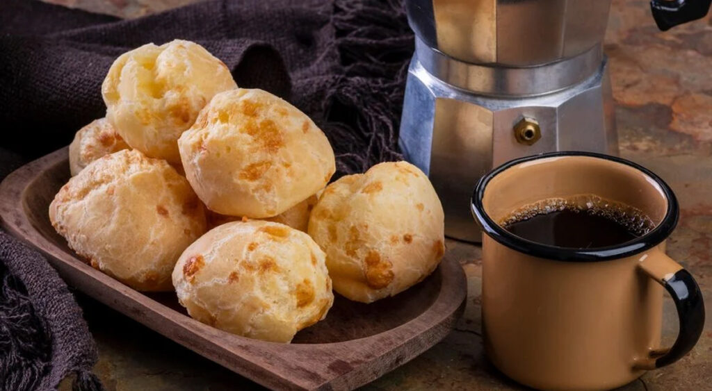 A imagem é uma foto que mostra uma bandeja de madeira com seis pães de queijo dourados, acompanhada por uma caneca bege com café preto e uma cafeteira de alumínio ao fundo.