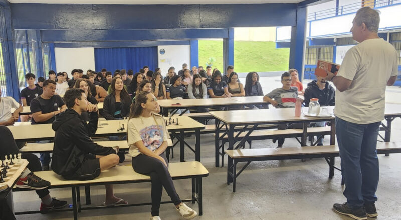 A imagem é uma foto do professor Eloésio Paulo falando para um grupo de estudantes da Escola Estadual Ismael Brasil Corrêa. O ambiente apresenta paredes pintadas de azul e áreas abertas que permitem a entrada de luz natural, mostrando grama ao fundo. Nas mesas à frente, há peças de xadrez e garrafas. A maioria dos alunos aparenta estar focada no que é apresentado.