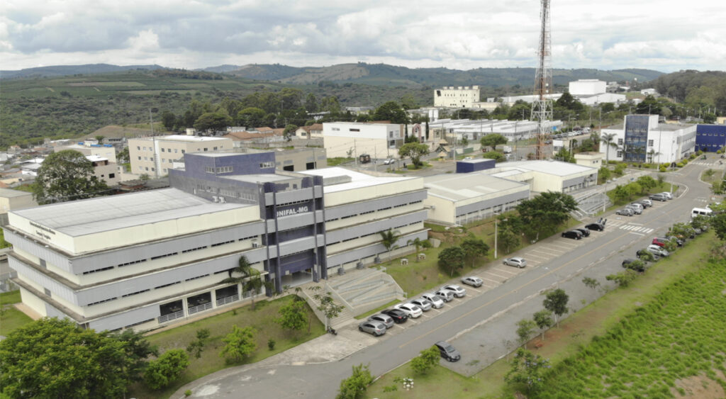 A imagem é uma foto aérea do campus Varginha da UNIFAL-MG, localizado em área verde e arborizada, com edifícios predominantemente de tom claro e detalhes em azul.