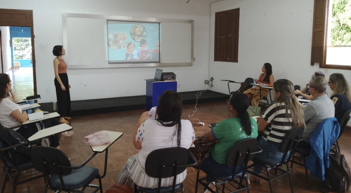 A imagem mostra uma foto tirada durante o curso de formação nas dependências da Secretaria Municipal de Educação de Alfenas, onde aparece um projetor, uma palestrante e os participantes sentados.