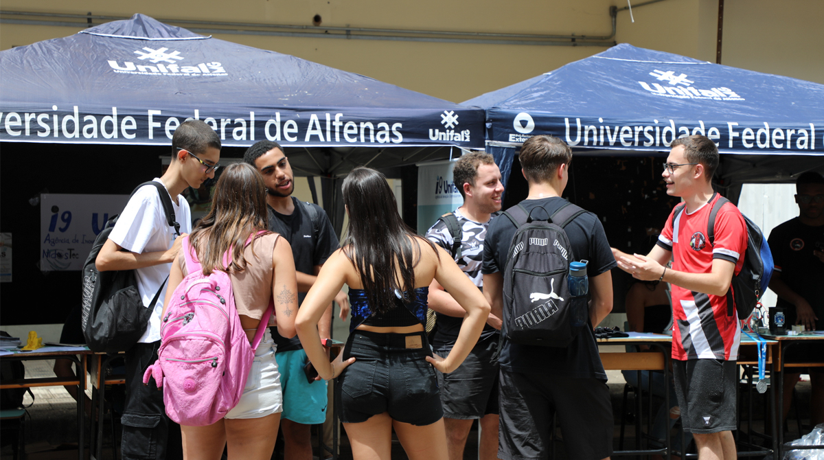 A imagem é uma foto de um grupo de estudantes da UNIFAL-MG conversando em pé, ao ar livre, em frente a tendas azuis com o logotipo e o texto "Universidade Federal de Alfenas".