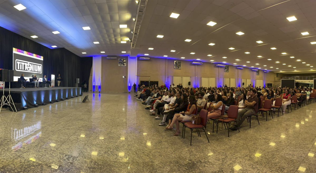 A imagem mostra a foto do salão do evento com fileiras de cadeiras preenchidas por dezenas de pessoas. À esquerda, há um palco elevado com quatro pessoas sentadas, participando de uma discussão ou painel.