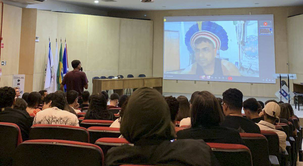 A imagem mostra a foto do auditório Leão de Faria com os alunos das escolas parceiras de Alfenas sentados em cadeiras vermelhas voltadas para a frente, onde um estudante em pé, de costas para a câmera, segura um microfone e se dirige ao telão à frente, onde o cacique Pawanã, da etnia Kariri-Xocó, aparece em uma chamada de vídeo.