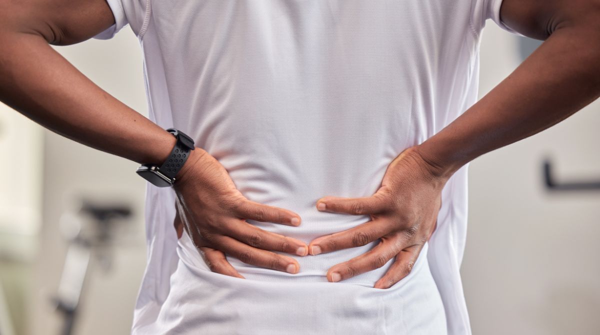 A imagem é uma foto de uma pessoa de costas, vestindo uma camiseta branca, com ambas as mãos apoiadas na região lombar. A posição das mãos e o corpo levemente curvado sugerem desconforto ou dor nas costas.