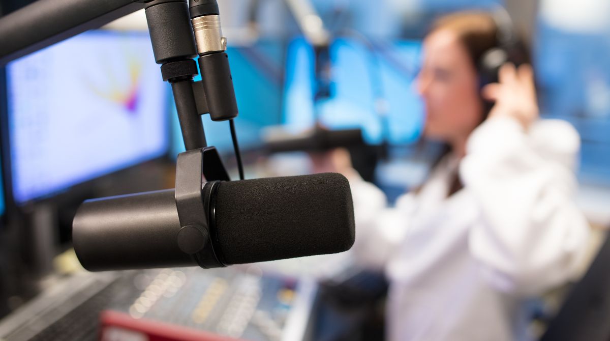 A imagem mostra um estúdio de rádio. Em primeiro plano, um microfone profissional preto com espuma protetora está fixado em um suporte articulado. Ao fundo, desfocado, uma pessoa com cabelos castanhos, vestindo uma roupa branca, ajusta os fones de ouvido enquanto está sentada diante de monitores de computador com luz azulada.