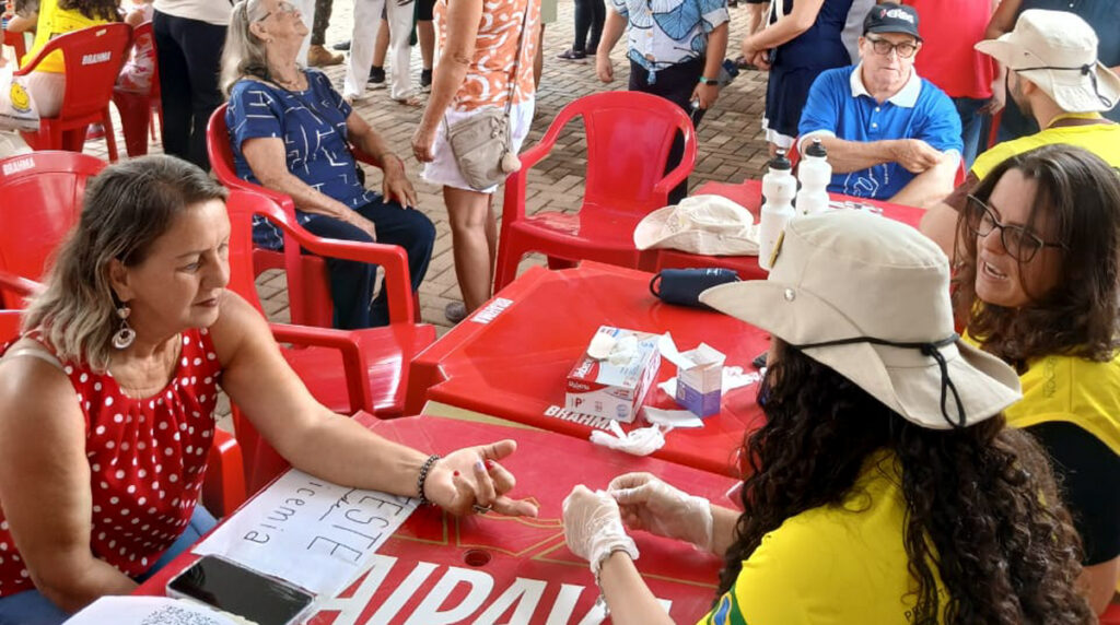 A imagem é uma foto dos rondonistas em atividade na cidade de Conceição dos Ouros em uma ação comunitária em um ambiente ao ar livre. No centro, uma mulher de blusa vermelha com bolinhas brancas está sentada à mesa, estendendo a mão para uma atendente com luvas, que realiza um teste de saúde.