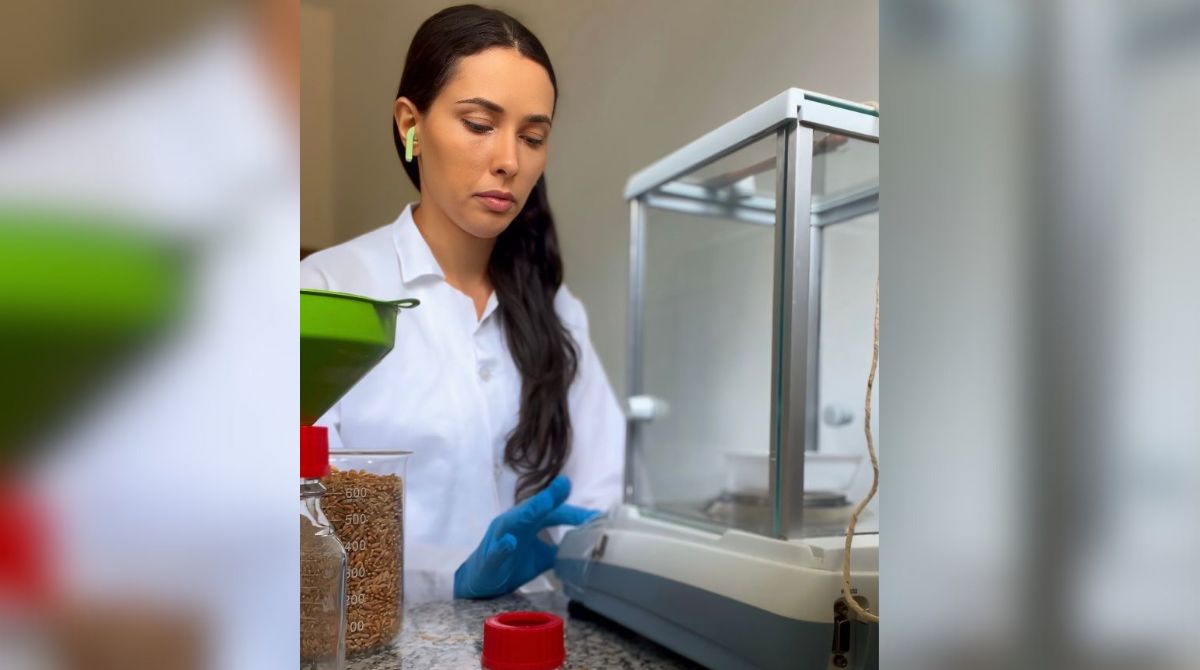 A imagem é uma foto da pesquisadora Surama Hualde em um laboratório de pesquisa. Ela veste um jaleco branco, luvas azuis e está em frente à bancada de granito.