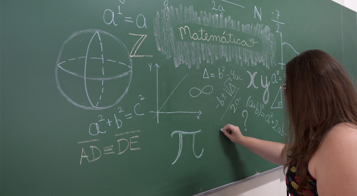 A imagem mostra uma foto da acadêmica Débora Perizato do curso de Matemática em frente a uma lousa verde repleta de fórmulas matemáticas e gráficos desenhados com giz branco e colorido.