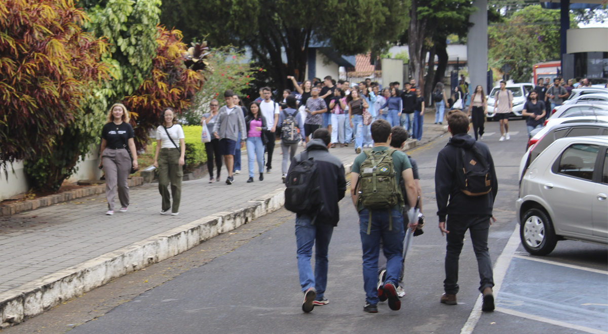 A imagem é uma foto feita no ambiente externo da Sede da UNIFAL-MG, em Alfenas, mostrando um grupo de pessoas caminhando por uma calçada e pela rua ao lado de um estacionamento com carros estacionados.