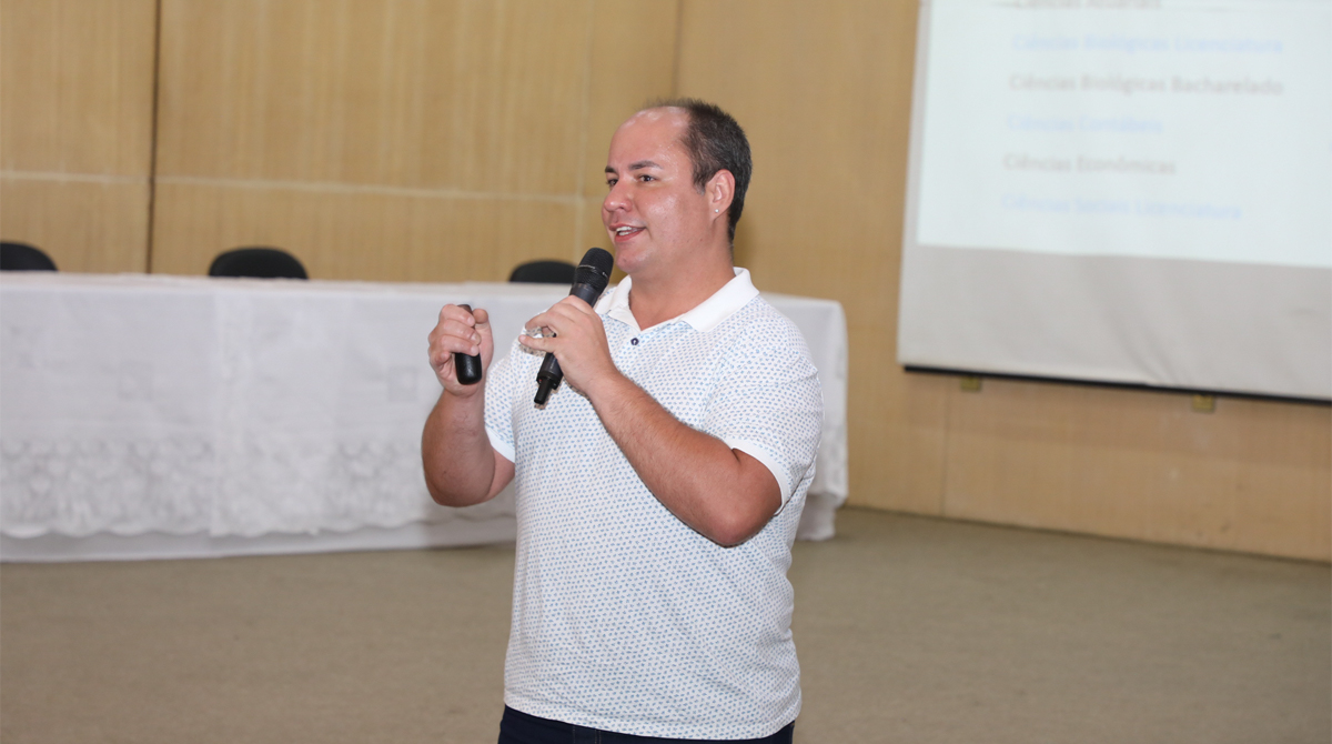 A imagem é uma foto do Geraldo Liska, que apresenta uma palestra em um auditório. Ele está em pé, segurando um microfone na mão direita e um controle remoto na mão esquerda. Ele sorri levemente enquanto fala. O ambiente sugere um evento acadêmico ou institucional.