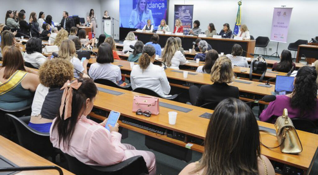 A imagem mostra a foto de uma sala de reuniões onde se encontra uma bancada formada por mulheres na parte central no fundo, e em frente a essa bancada outras fileiras ocupadas por mulheres de costas.