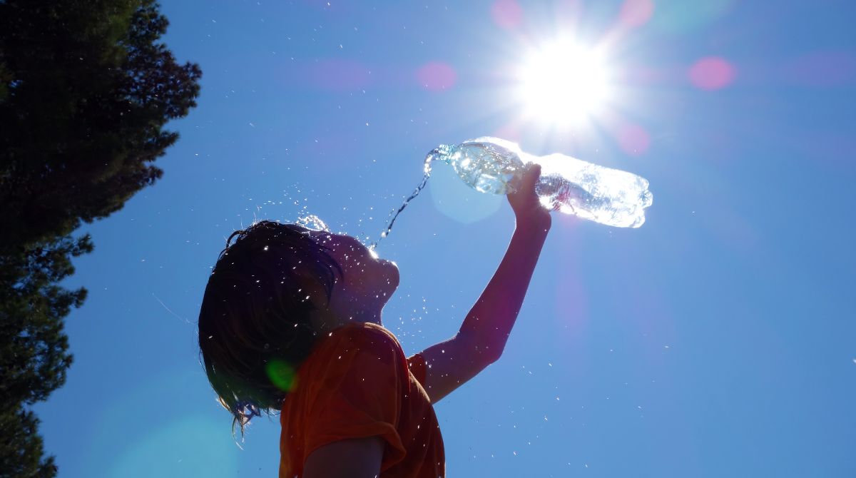 A imagem mostra a silhueta de uma criança vestindo camiseta laranja, inclinada para trás enquanto bebe água de uma garrafa plástica transparente. O sol brilha forte no céu azul, criando reflexos na água e iluminando as gotículas que caem ao redor.