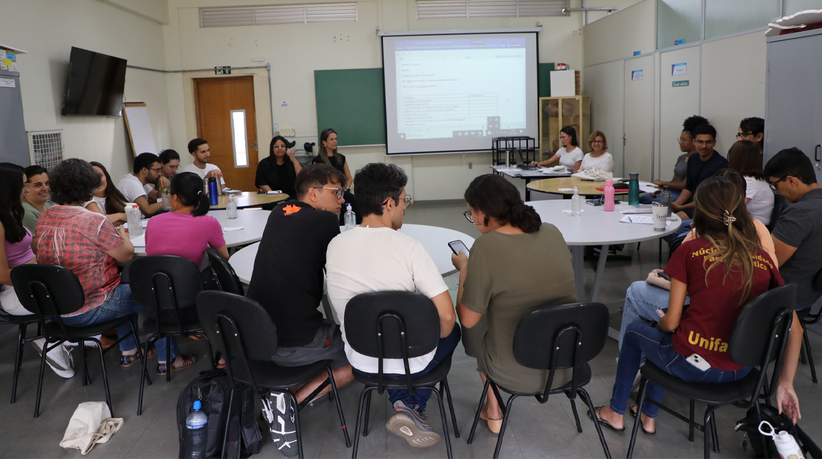 A imagem mostra uma foto do grupo de estudantes e profissionais de saúde reunidos em uma sala com paredes claras, projetor ligado e quadro verde ao fundo. Cerca de vinte pessoas, em sua maioria jovens, estão sentadas em cadeiras pretas ao redor de mesas redondas, participando de uma discussão.