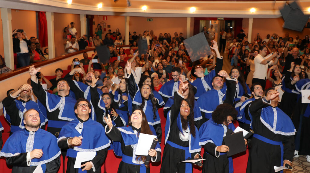 A foto mostra os formandos em becas azul e preta, jogando os capelos para o alto no auditório do Theatro Capitólio, em Varginha.