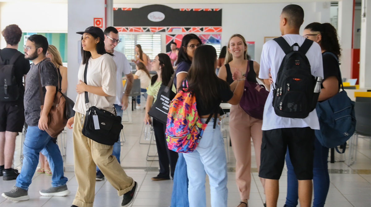 A imagem é uma foto de um grupo de estudantes caminhando e interagindo na área da cantina do prédio V da Sede da UNIFAL-MG.