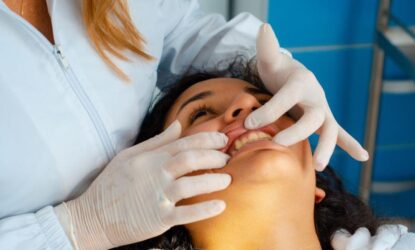 Imagem mostra uma foto em close com uma paciente deitada em uma cadeira odontológica, sorrindo, enquanto uma dentista com luvas de látex brancas examina seus dentes com as mãos.